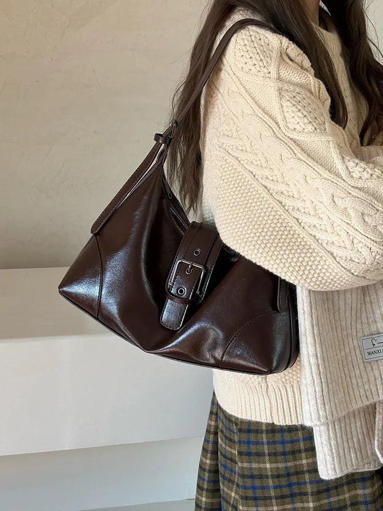 A brown retro PU leather underarm bag carried by a woman wearing a cream knit sweater and plaid skirt.