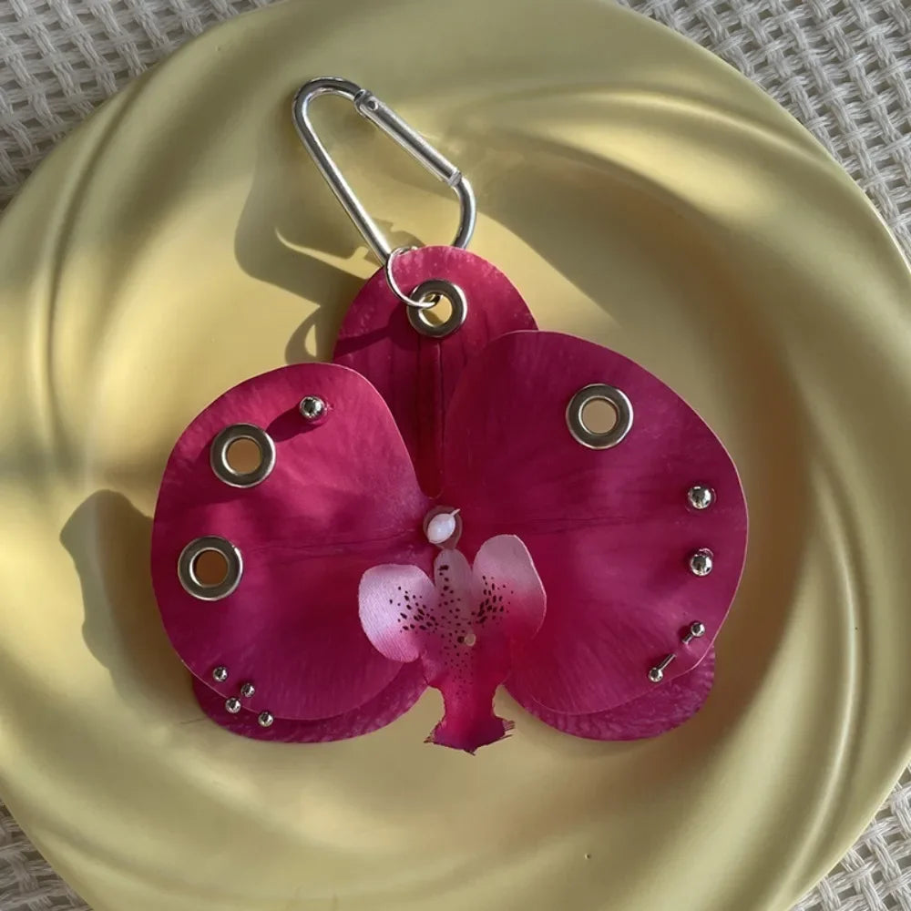 A realistic magenta orchid flower made of fabric is attached to a silver keychain, resting on a gold plate.