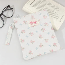 A white floral plaid cotton tablet pouch with a pink tag and a drawstring is shown next to eyeglasses and a book.