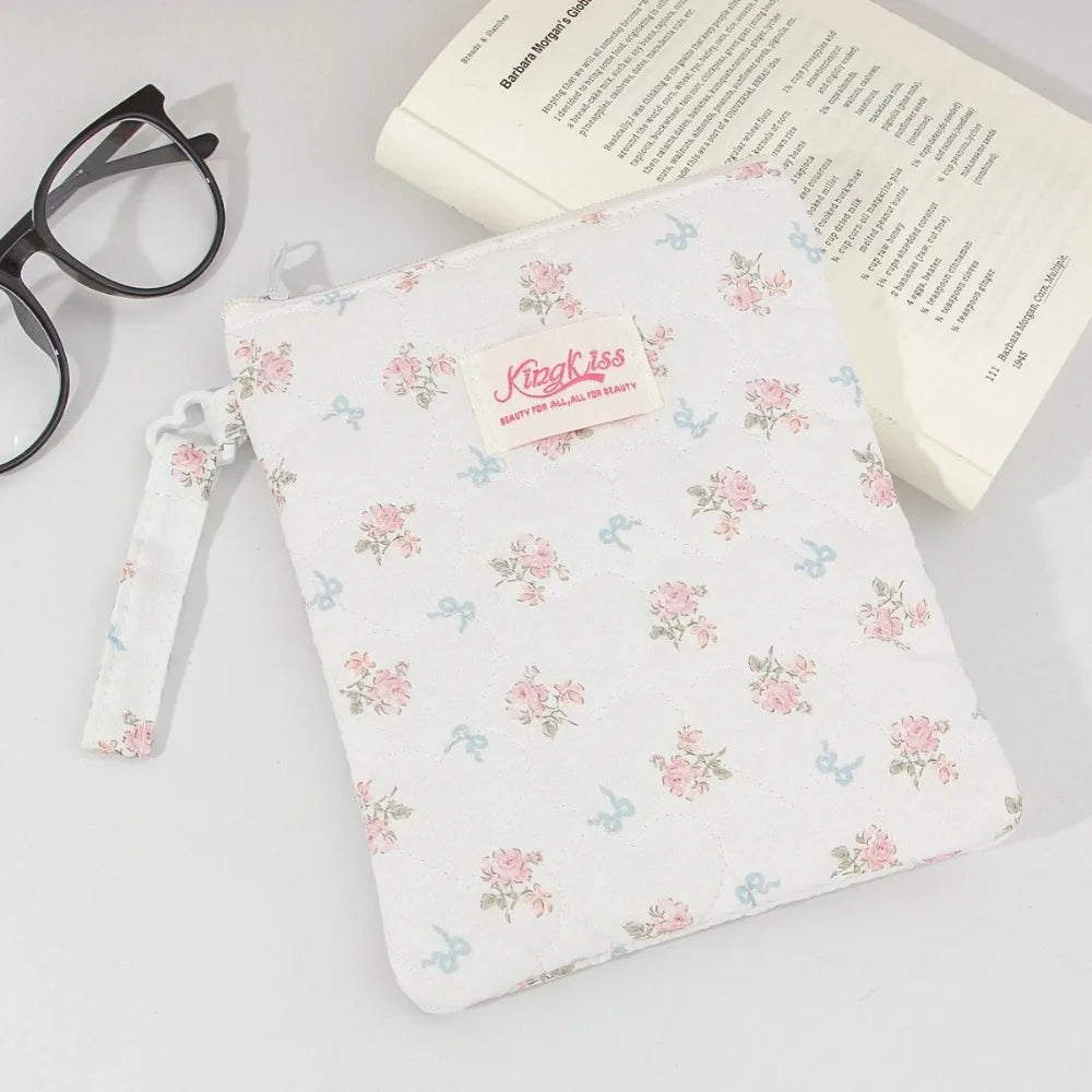 A white floral plaid cotton tablet pouch with a pink tag and a drawstring is shown next to eyeglasses and a book.