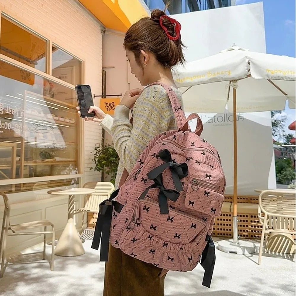 A woman wearing a light yellow sweater and brown skirt holds a pink corduroy bow print backpack outdoors.