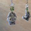 A small, gray felted creature pendant with large eyes and a green bead charm on a silver chain.