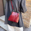 A woman is holding a red leather shell handbag with a strap against a brick wall.