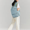 A woman wearing a beige cap and white shirt carries a light blue backpack while standing.