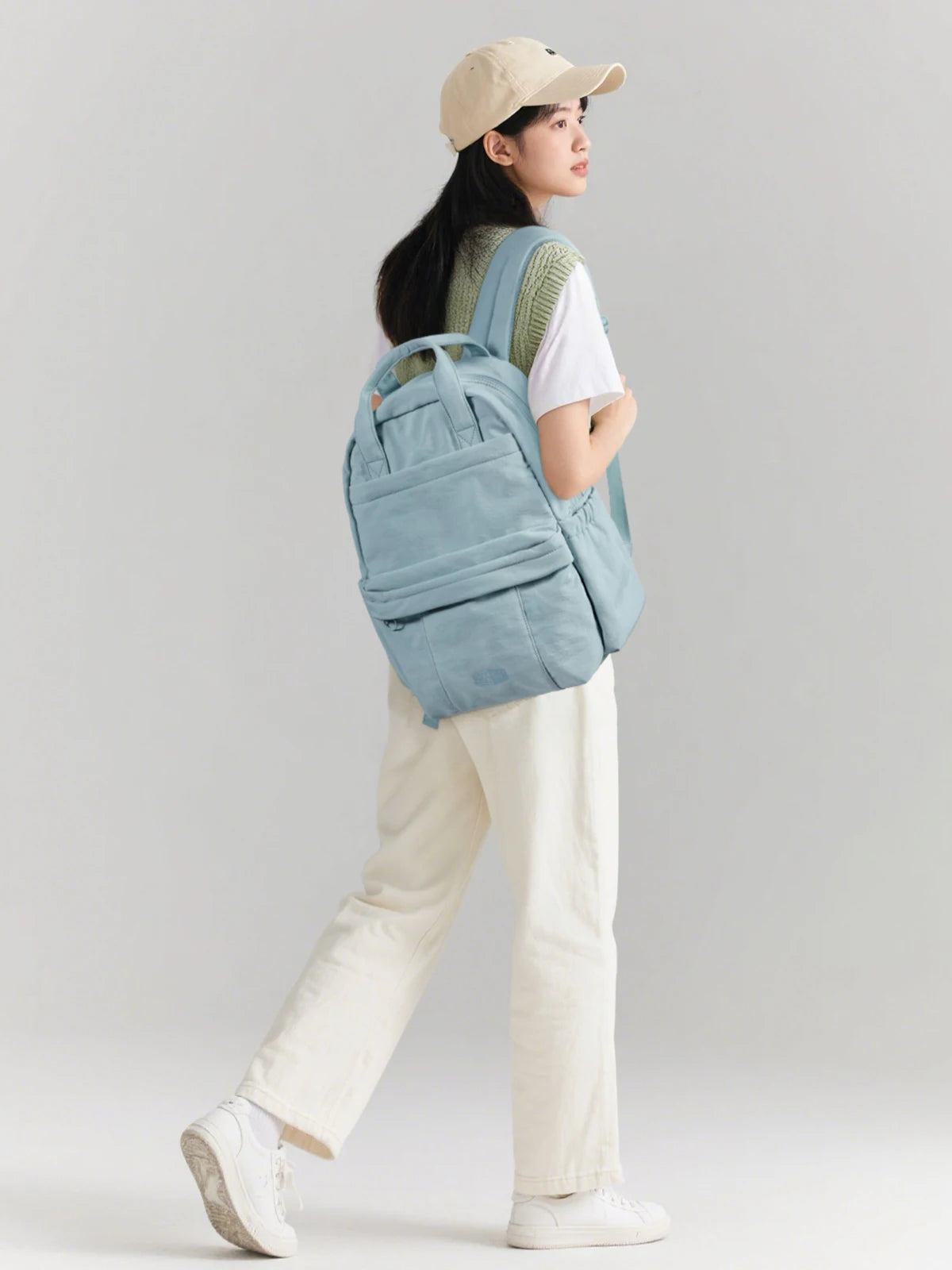 A woman wearing a beige cap and white shirt carries a light blue backpack while standing.