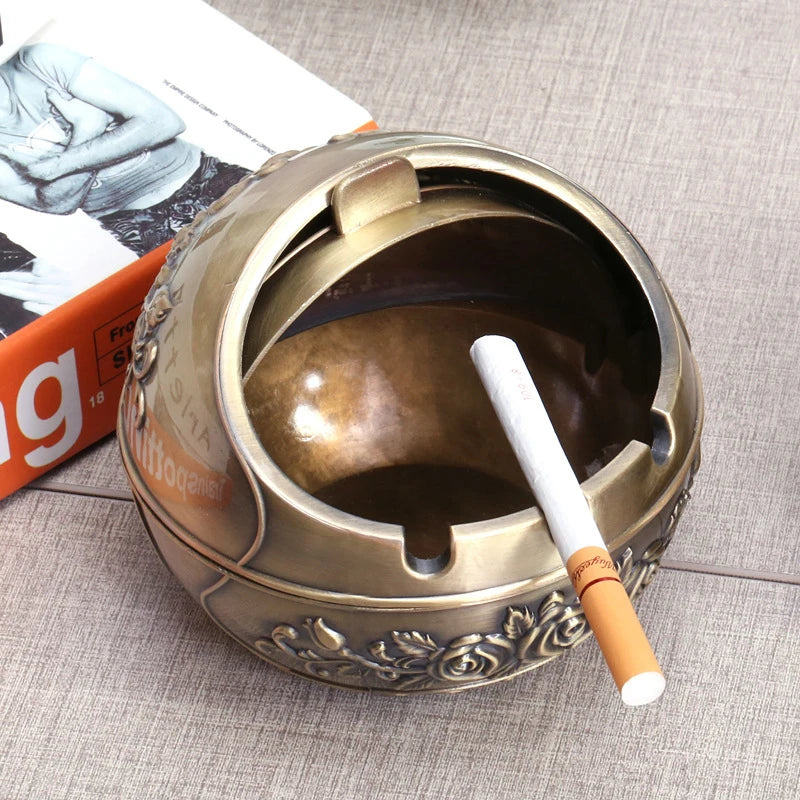 Vintage metal ashtray with a rose embossing and windproof lid resting on a light gray surface.