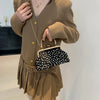 A woman wearing a camel-colored blazer and skirt holds a black studded crossbody bag with white pearls.