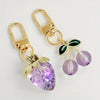 Gold-toned keychain set featuring a purple fruit charm and a green leaf charm.