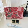 A pink floral patterned cosmetic pouch next to a gold square and a makeup brush.