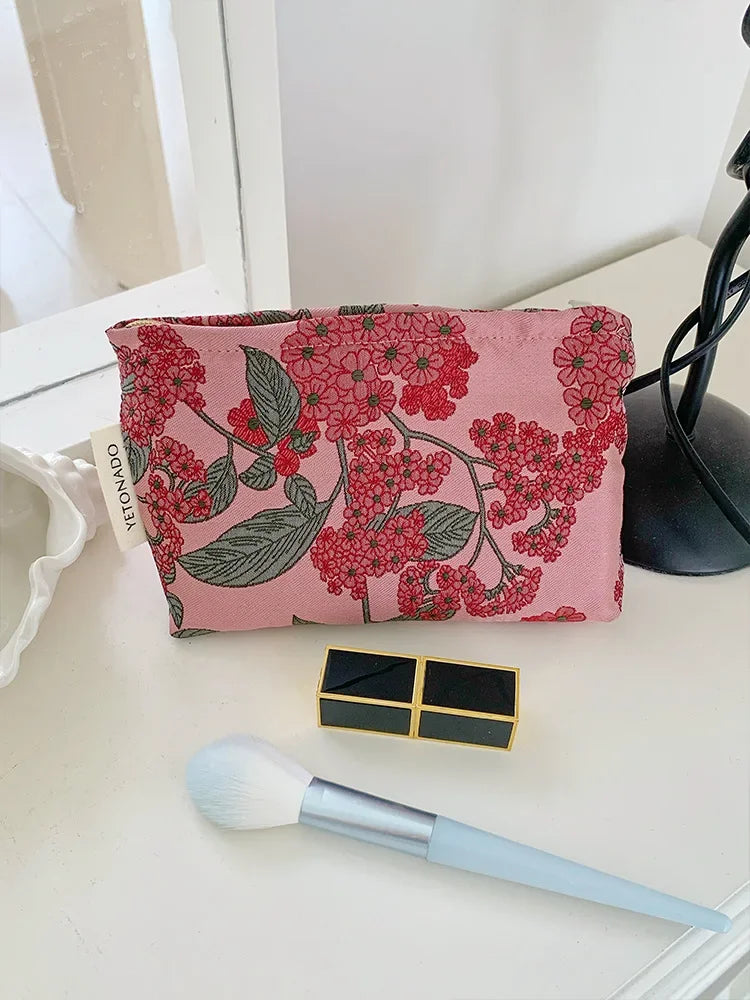 A pink floral patterned cosmetic pouch next to a gold square and a makeup brush.