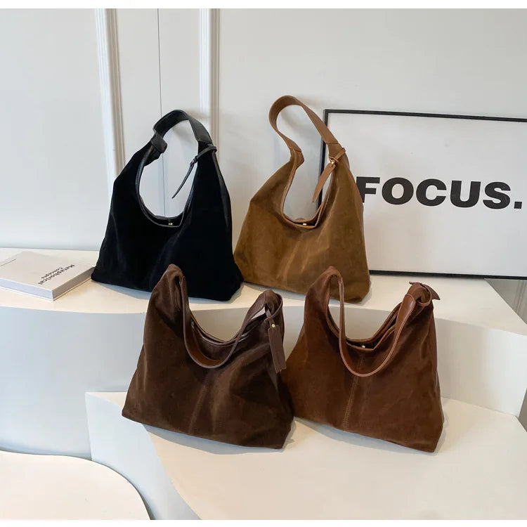 Three suede slouchy shoulder bags in black and brown resting on a white surface with a black framed sign.