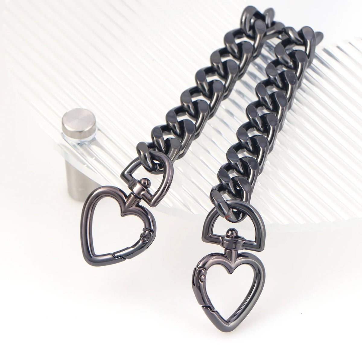 A dark metal heart-shaped chain extender is displayed against a clear, ribbed background.