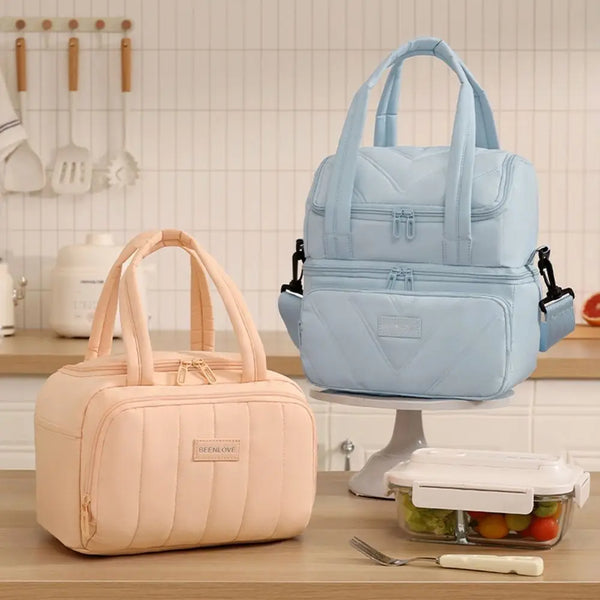 A light blue reusable insulated lunch bag is displayed on a stand next to a pink version.