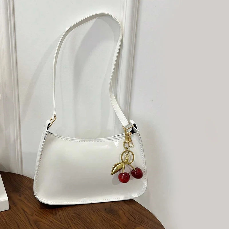 A white PU leather shoulder bag with a gold chain and cherry pendant resting on a wooden surface.