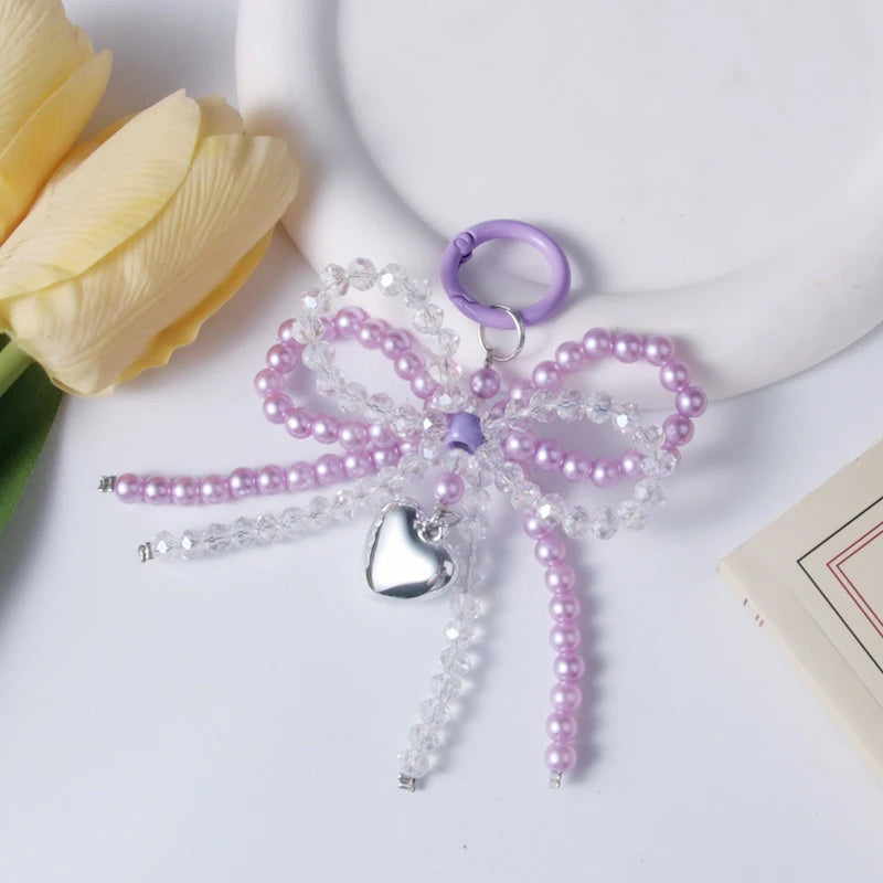 A purple and clear pearl bowknot keychain with a silver heart charm resting on a white surface next to a yellow rose.