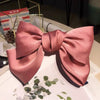 A satin pink bow hair clip is displayed on a white surface with green foliage.