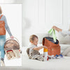 A woman wears a brown backpack diaper bag while sitting on a bed with a baby nearby.