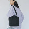 A woman wearing a white shirt holds a black canvas crossbody bag with an adjustable strap and multiple pockets.