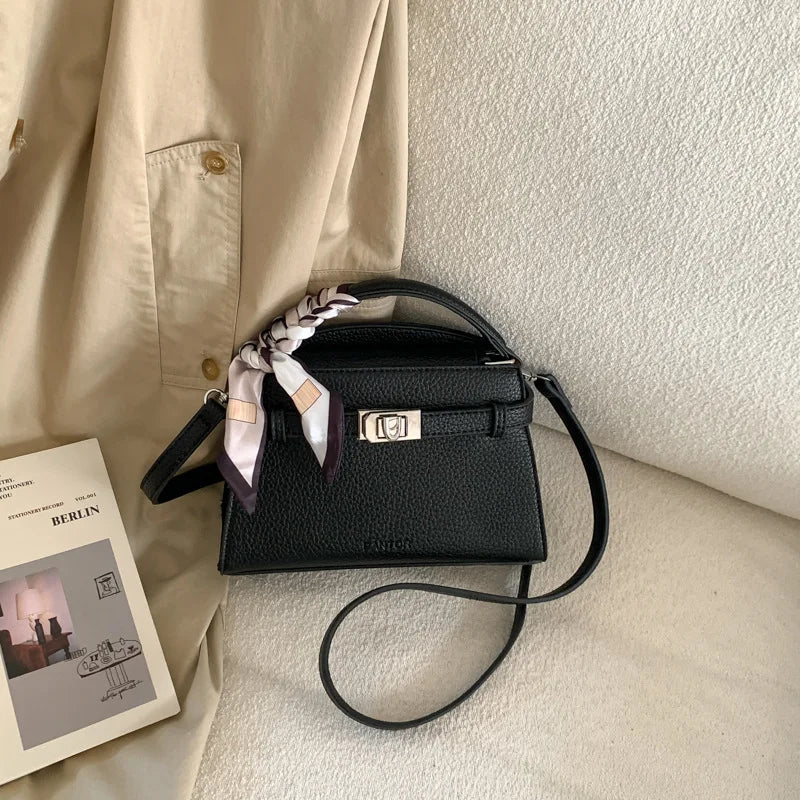 A black PU leather crossbody clutch bag with light pink and white striped fabric detail resting on a cream-colored garment.