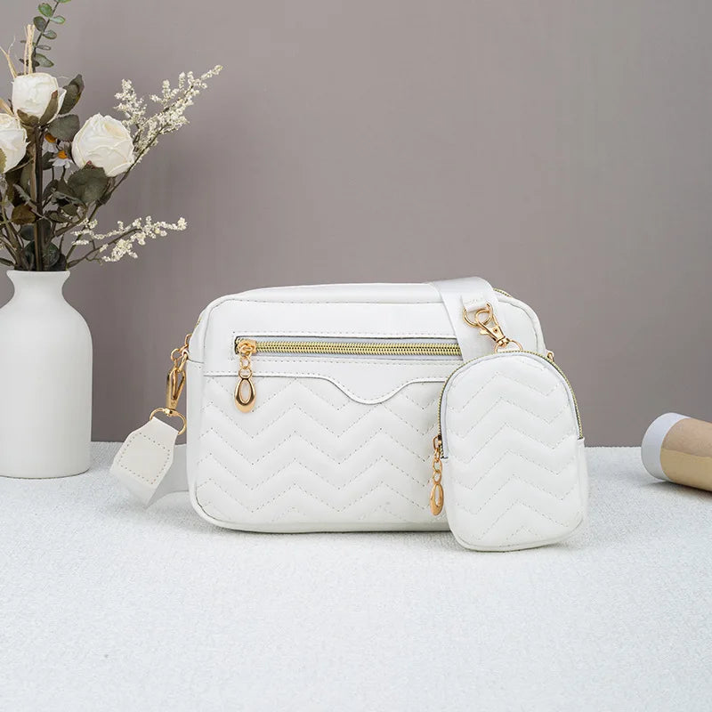 Weiße gesteppte Crossbodytasche mit goldfarbenen Details auf weißem Untergrund neben getrockneten Blumen.
