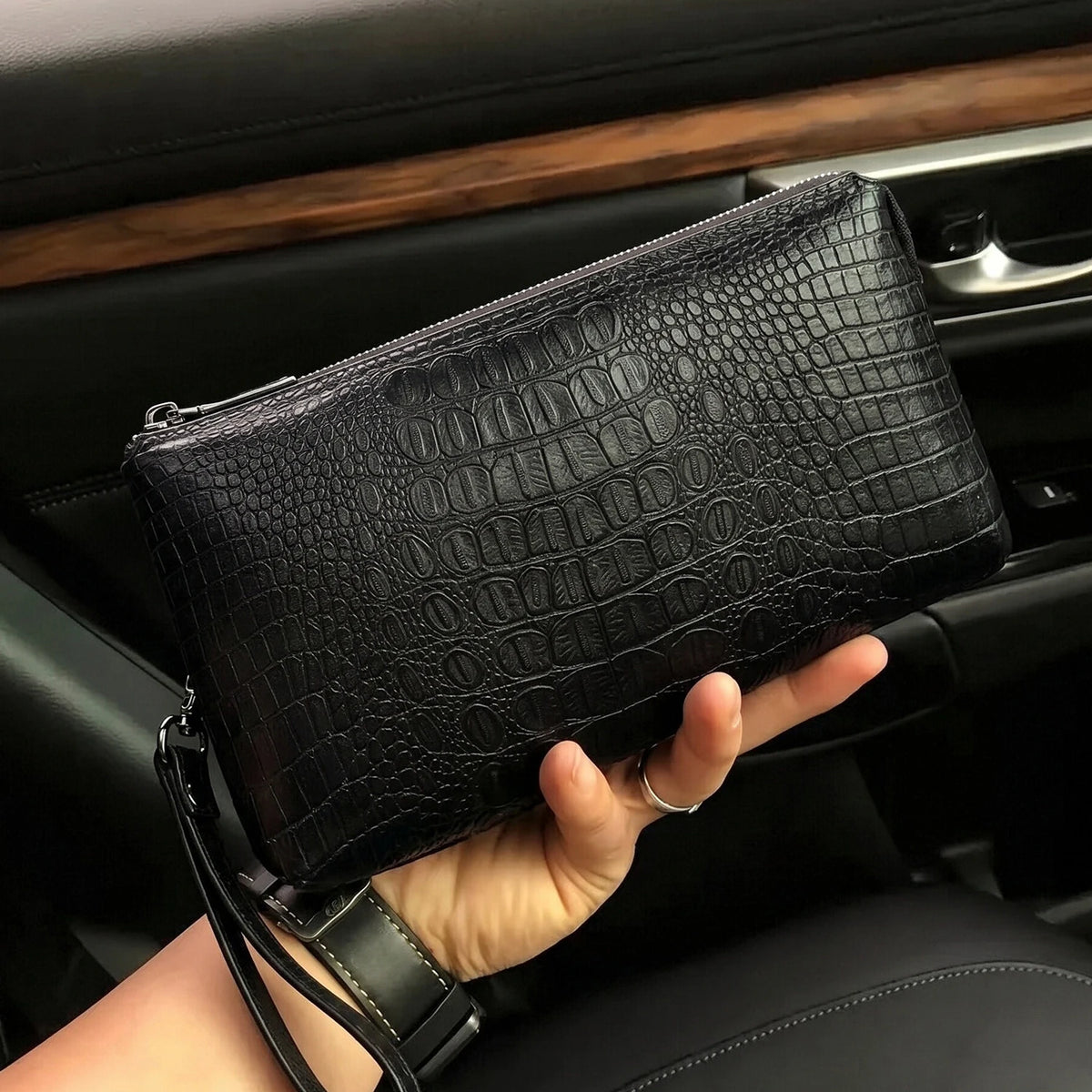 Un bolso de mano con estampado de cocodrilo negro sostenido en la mano de una persona dentro del interior de un coche.