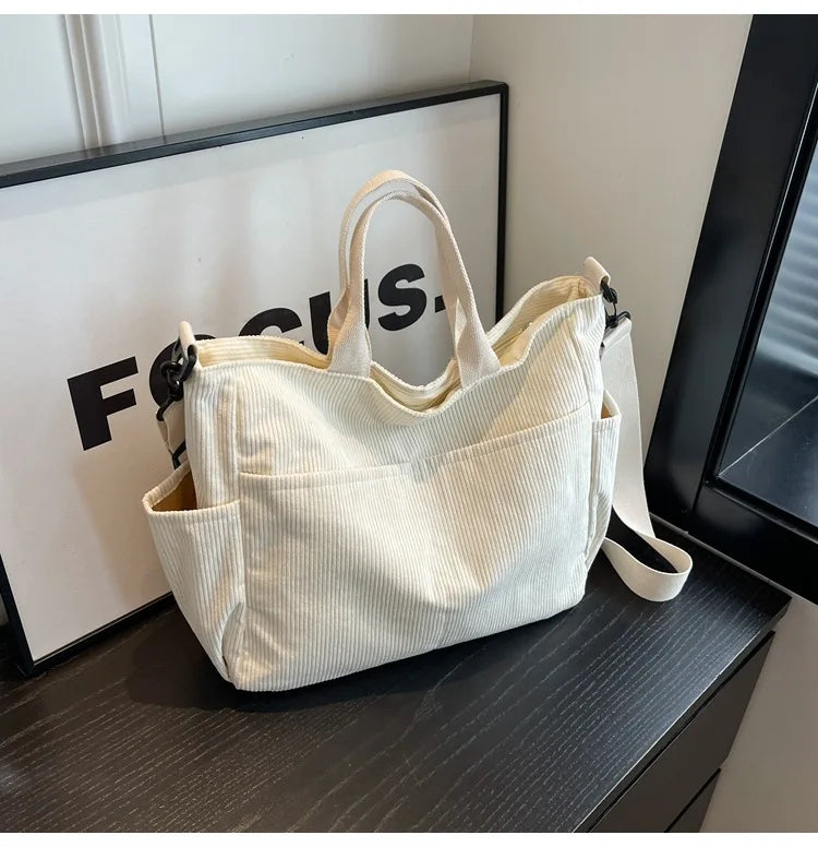 A cream corduroy multi-pocket tote bag with light vertical ribbing is displayed on a dark surface.