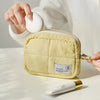 A light yellow quilted puffer makeup bag is shown being held by a hand on a white surface.