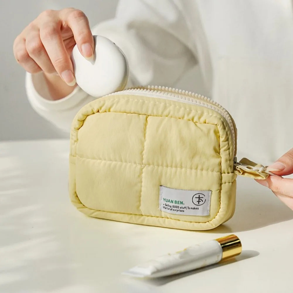 A light yellow quilted puffer makeup bag is shown being held by a hand on a white surface.