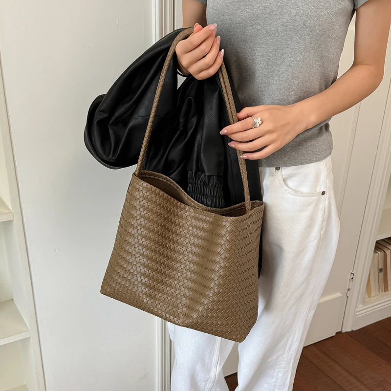 A person holds a brown woven bucket bag and a black leather garment against a white door frame.