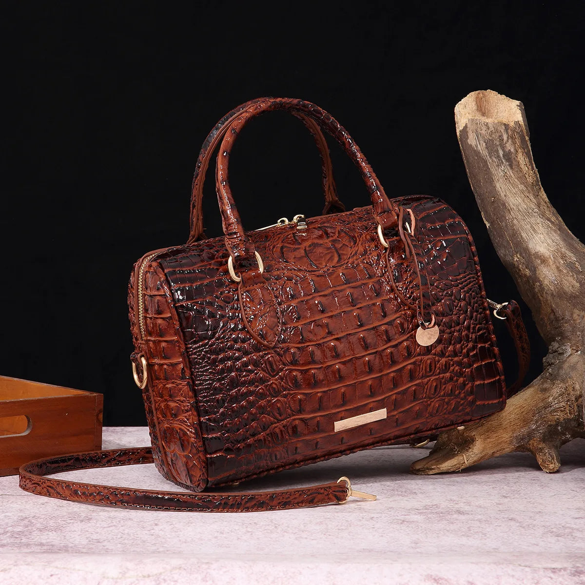 A brown crocodile pattern leather crossbody tote bag resting on a light surface next to a piece of driftwood.