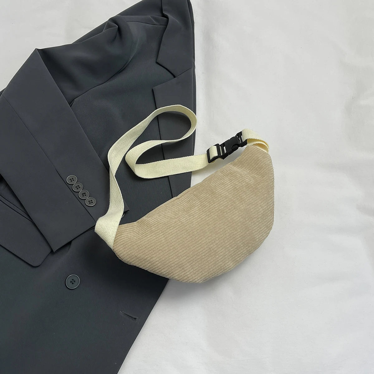 A beige corduroy crossbody waist bag with a light strap is shown next to a dark gray blazer.