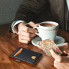 A hand holds a brown coffee cup and a clear RFID-blocking wallet on a wooden table.