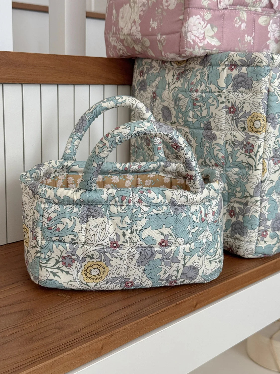 A quilted fabric storage basket in soft blue and cream floral patterns sits on a wooden shelf.
