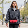 A woman wearing a dark jacket and jeans holds a red multi-pocket waist bag on a city street.