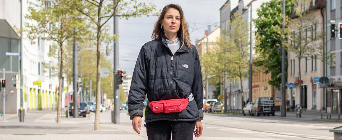 A woman wearing a dark jacket and jeans holds a red multi-pocket waist bag on a city street.