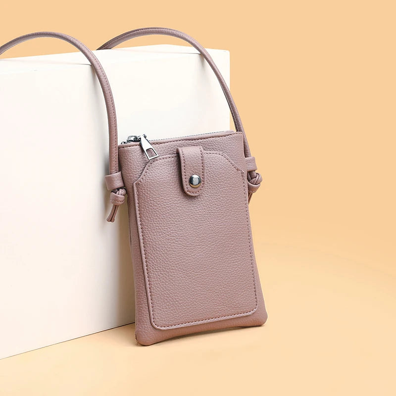 A light mauve compact PU leather crossbody bag with a zipper and leather strap resting on a beige surface.