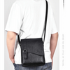 A man wearing a white t-shirt is holding a black PU leather messenger bag with an adjustable strap.