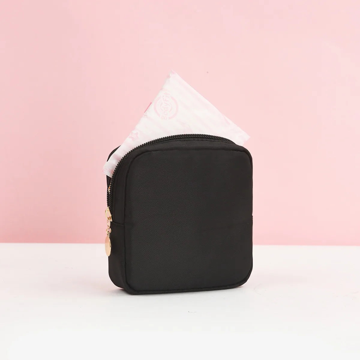 Schwarze Tragetasche für Damenbinden mit weißem Papiertupfer, vor rosa Hintergrund.