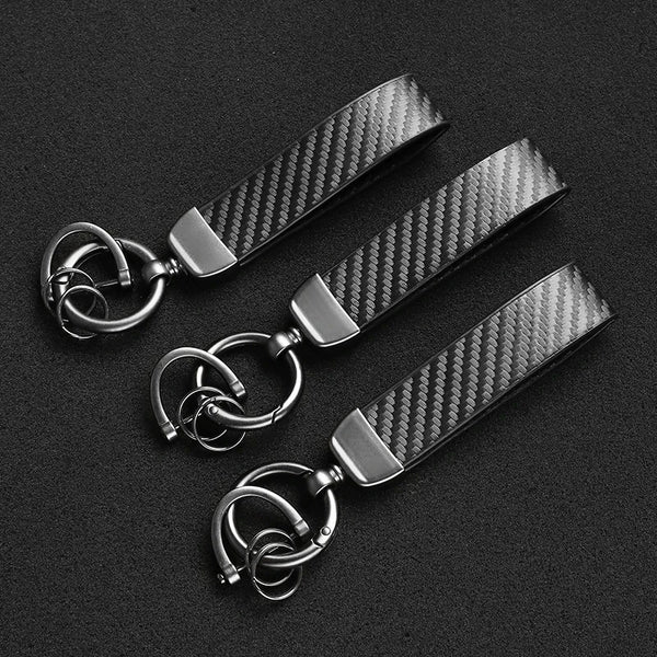 A black carbon fiber leather keychain with silver hardware lying on a dark surface.