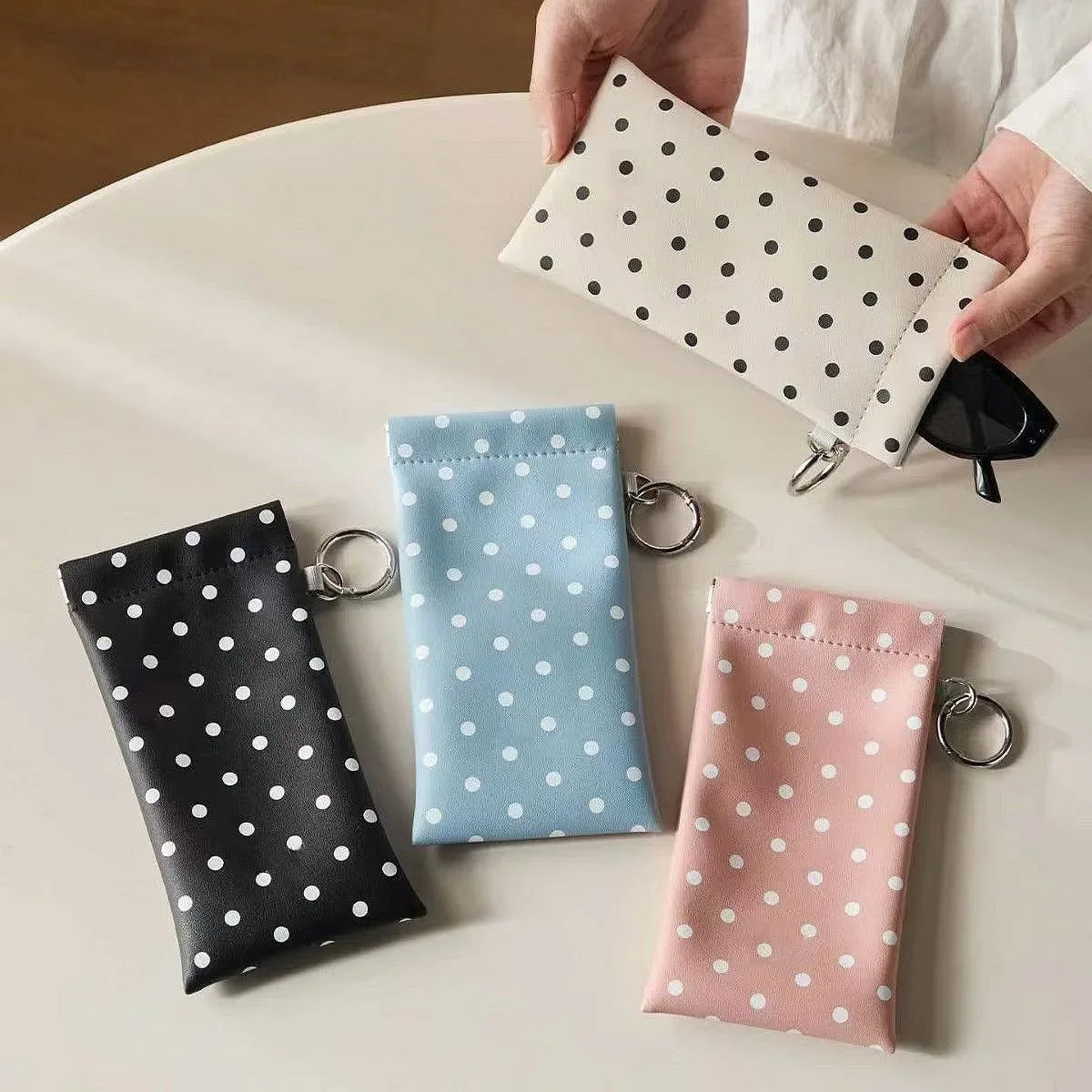 A collection of polka dot eyeglass pouches in black, white, blue, and pink, with a hand holding one pouch and a keychain.
