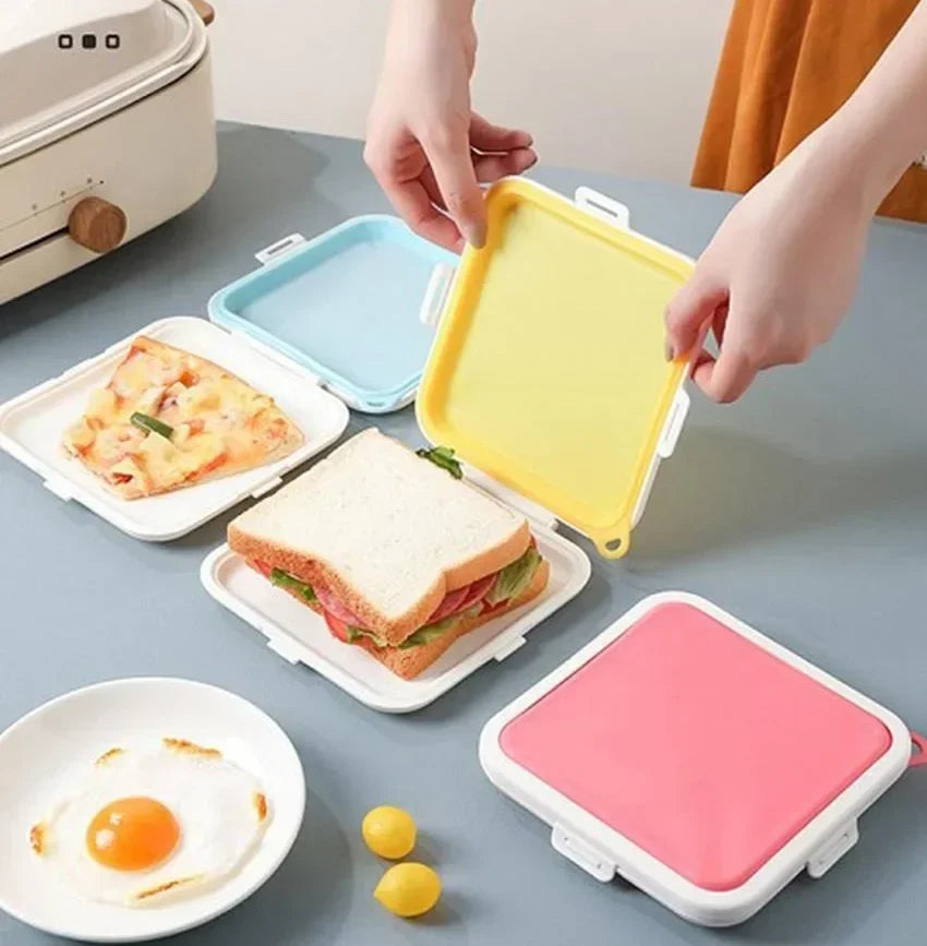 Hands are holding a yellow silicone reusable sandwich and lunch container next to a plate of food and a pink container.