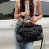 A woman holds a black saddle-shaped PU leather city tote bag in front of a white car.