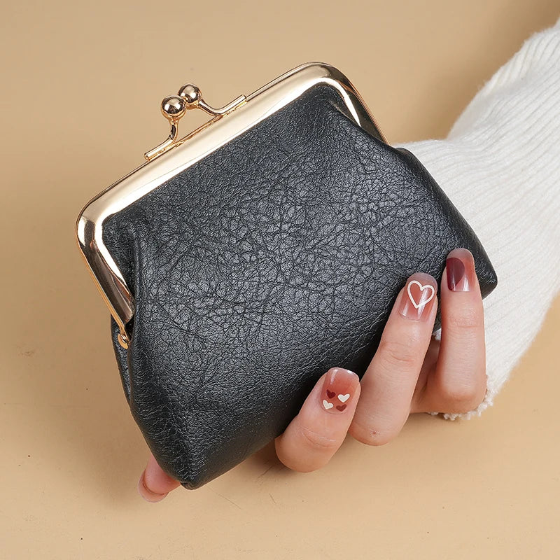 A black faux leather coin purse with a gold buckle is held by a hand against a beige background.
