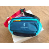 A teal and blue patchwork color block fanny pack with a red strap resting on a white box.