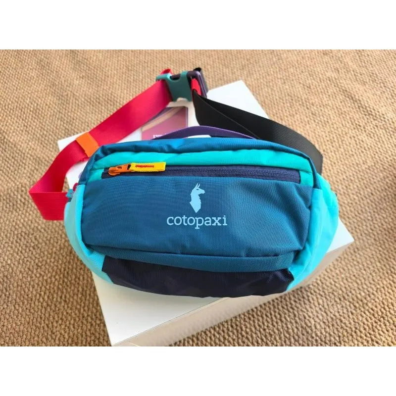 A teal and blue patchwork color block fanny pack with a red strap resting on a white box.