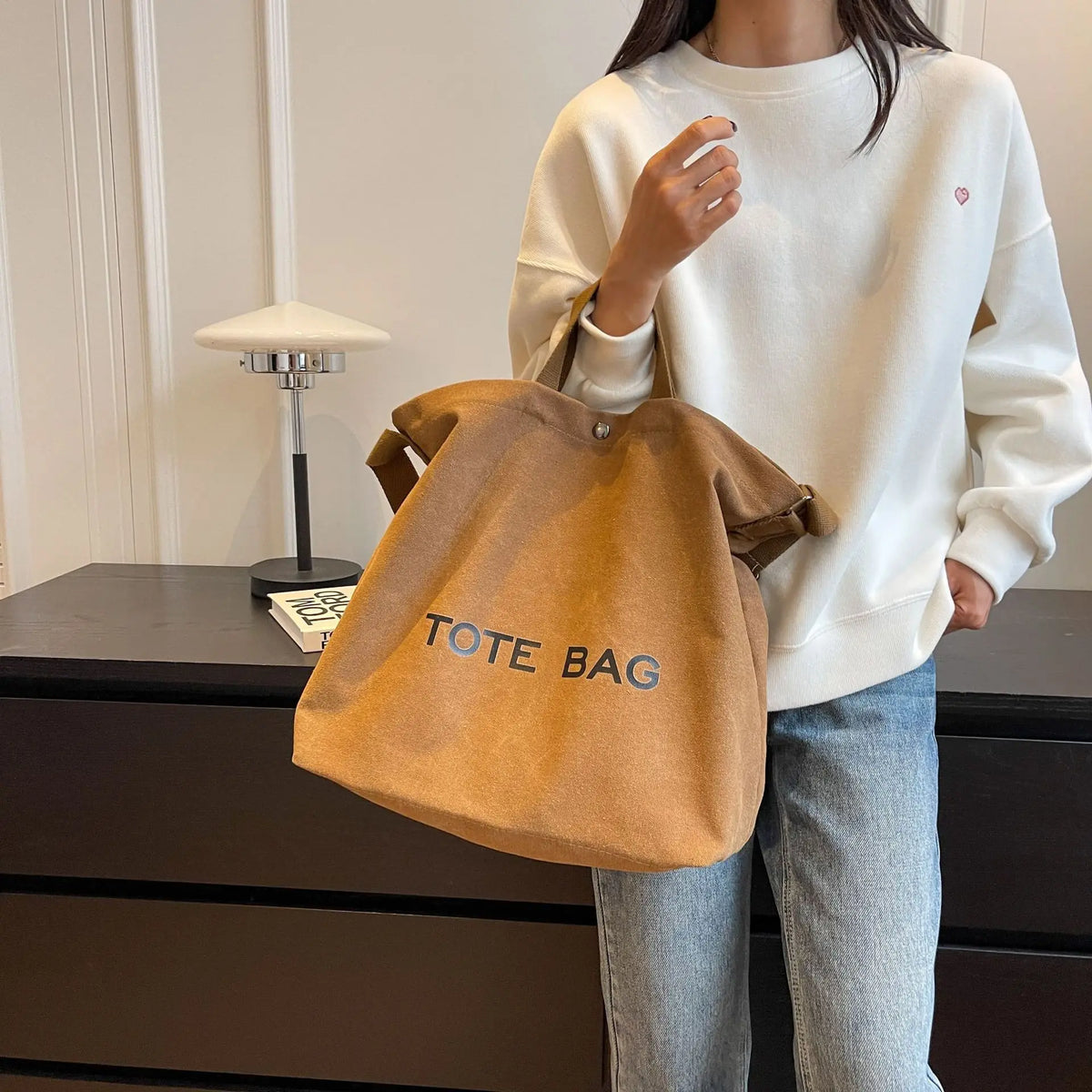 A woman wearing a white sweater holds an orange canvas tote bag in front of a dark cabinet.