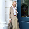 A woman wearing a beige trench coat and trousers stands next to a blue door frame holding a brown leather bucket bag.