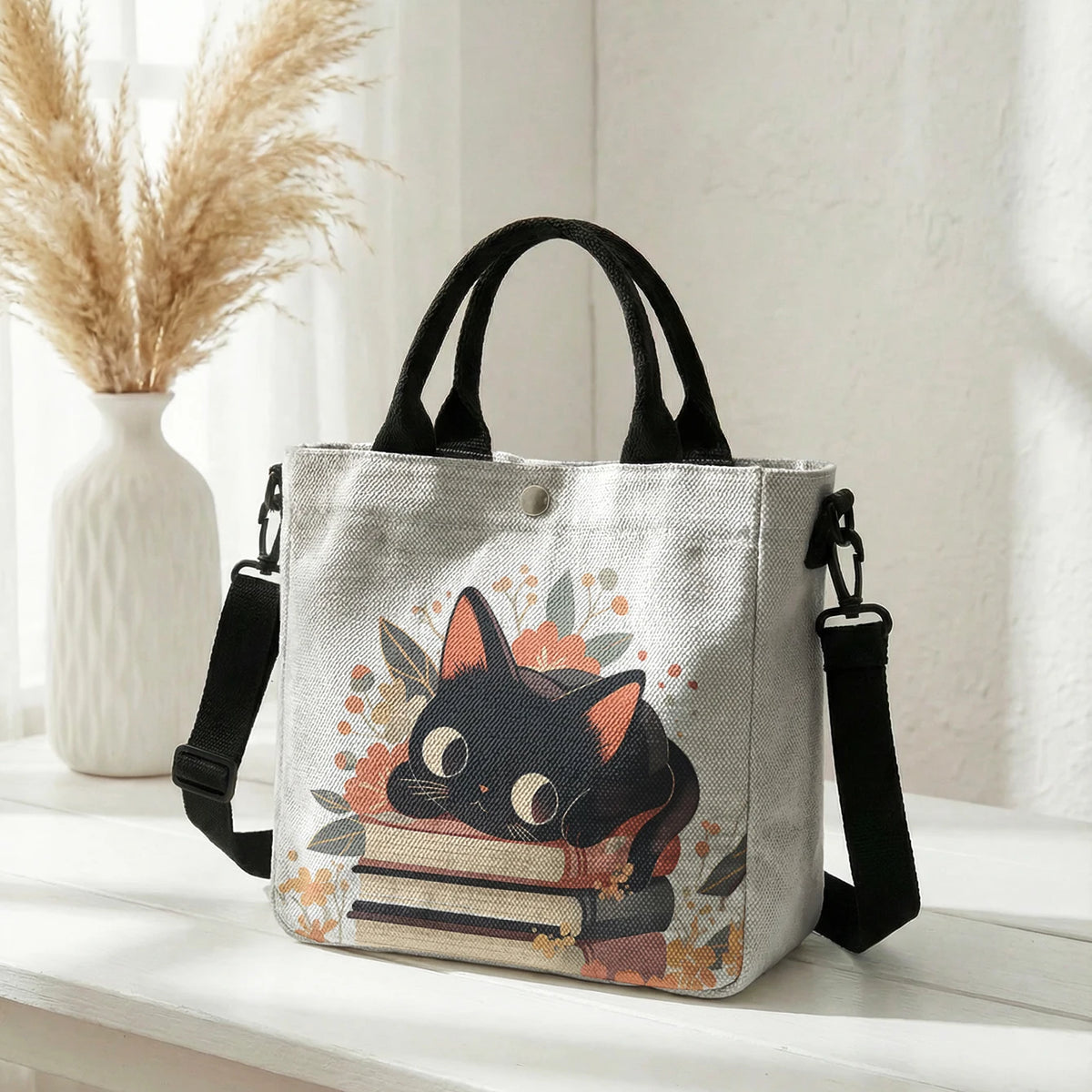 Graue Einkaufstasche mit Blumen-Katzenmuster, schwarzes Katzengesicht und offenes Buch, auf weißem Untergrund.