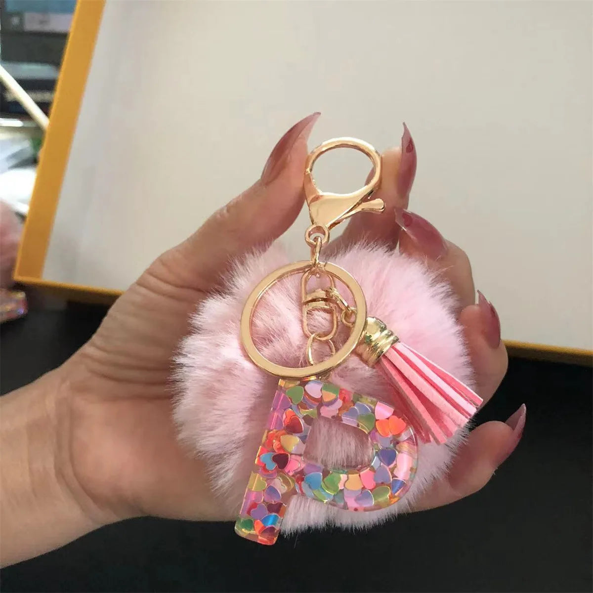 A hand holds a custom initial letter keychain featuring a shiny resin pendant and pink alphabet charm surrounded by fluffy pi