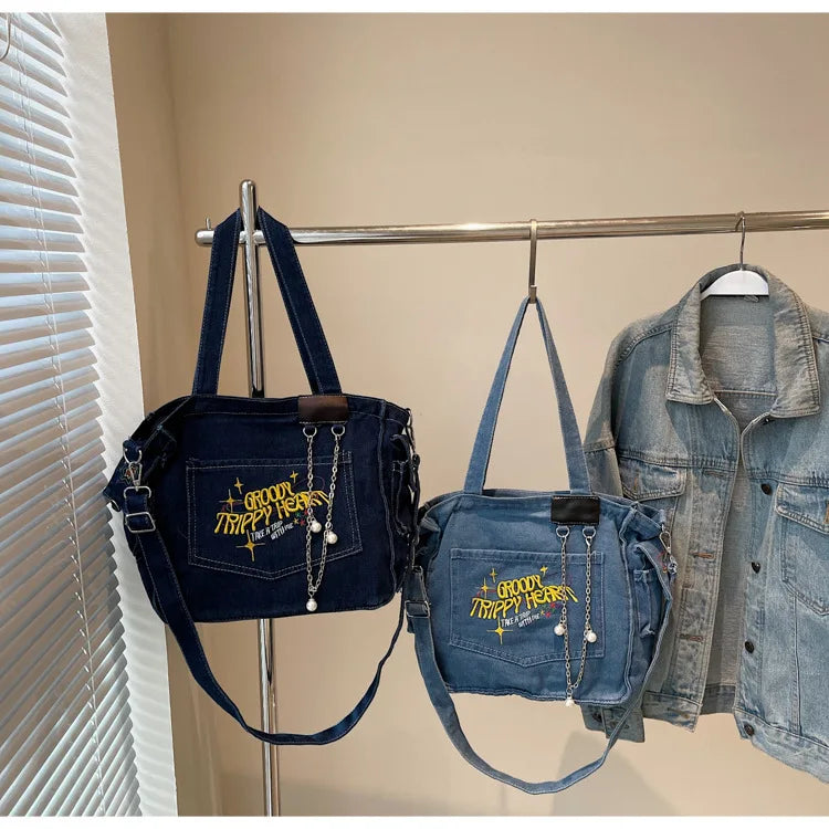 A navy blue denim crossbody bag is displayed on a hanger next to a denim shirt.
